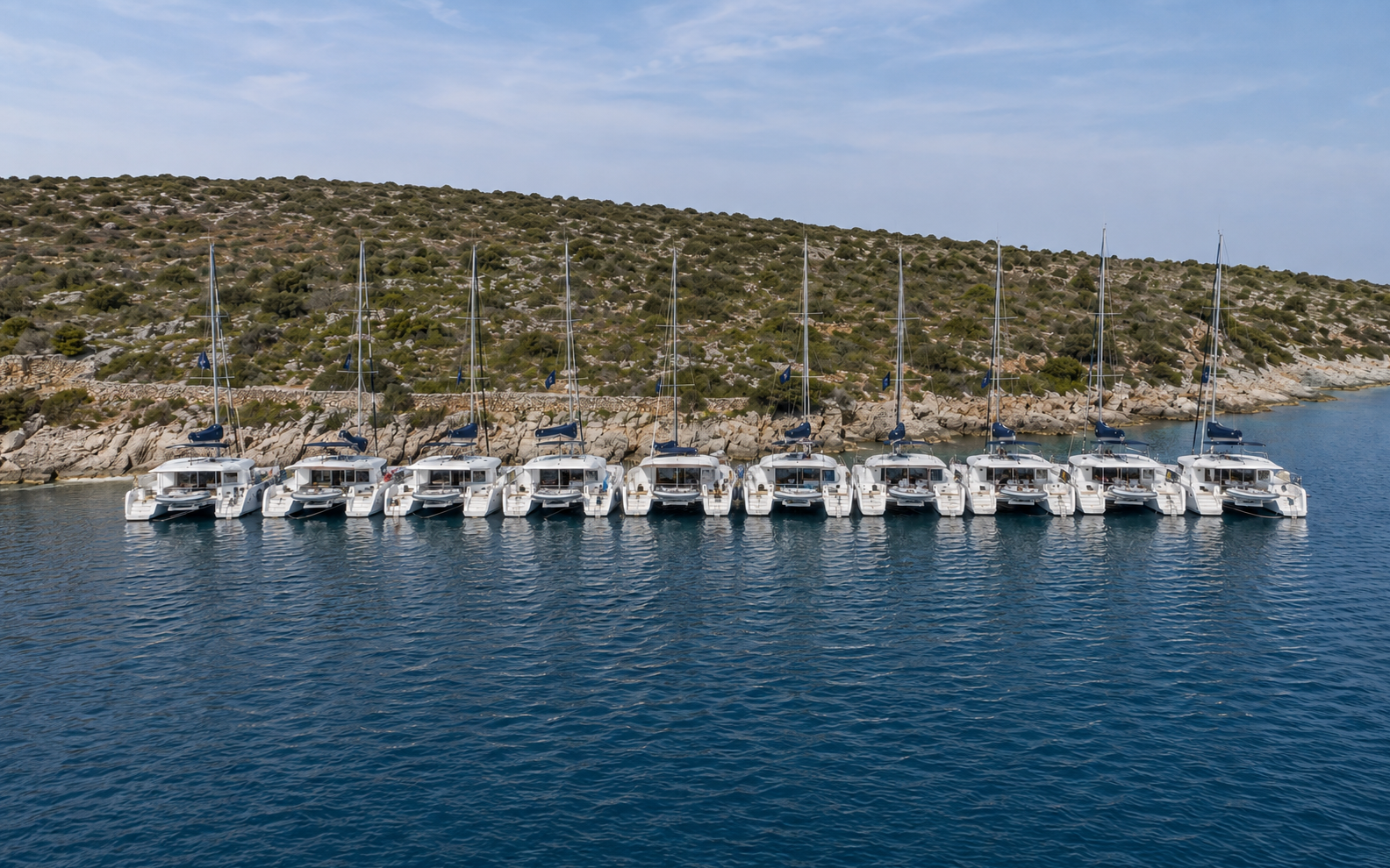 Aerial view of a fleet of identical charter catamarans moored side by side along a rocky Mediterranean shoreline.