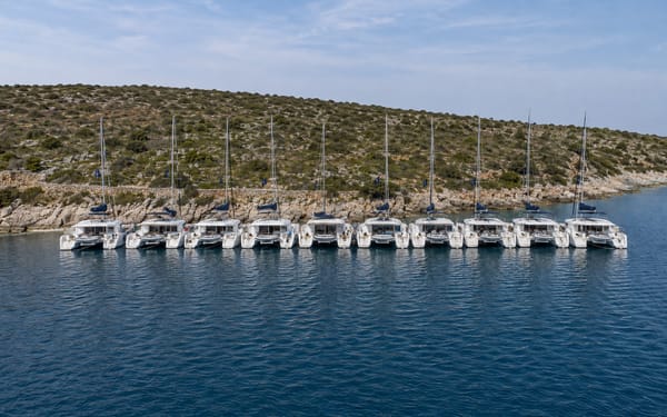 Aerial view of a fleet of identical charter catamarans moored side by side along a rocky Mediterranean shoreline.